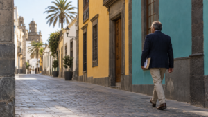 Hombre de mediana edad caminando por una calle de casas de colores en Canarias con un sobre en la mano, camino de resolver su revisión de incapacidad permanente